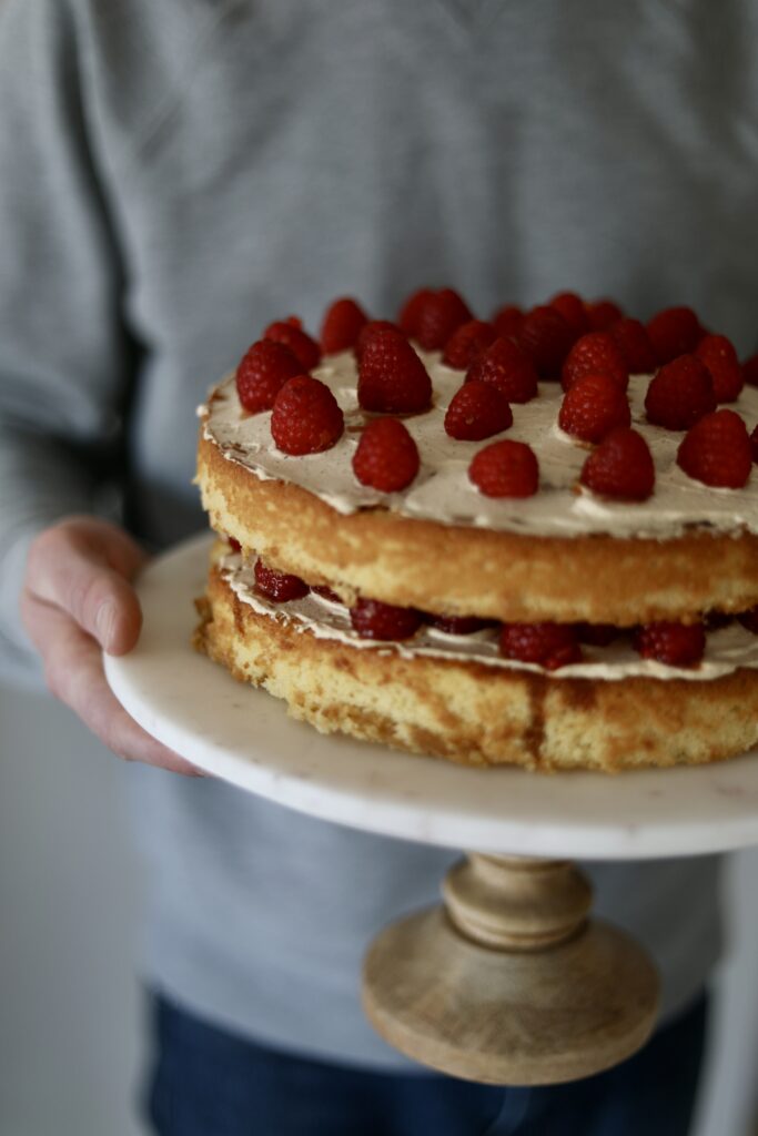 yellow cake with raspberries on top placed on a cake stand with hands holding it